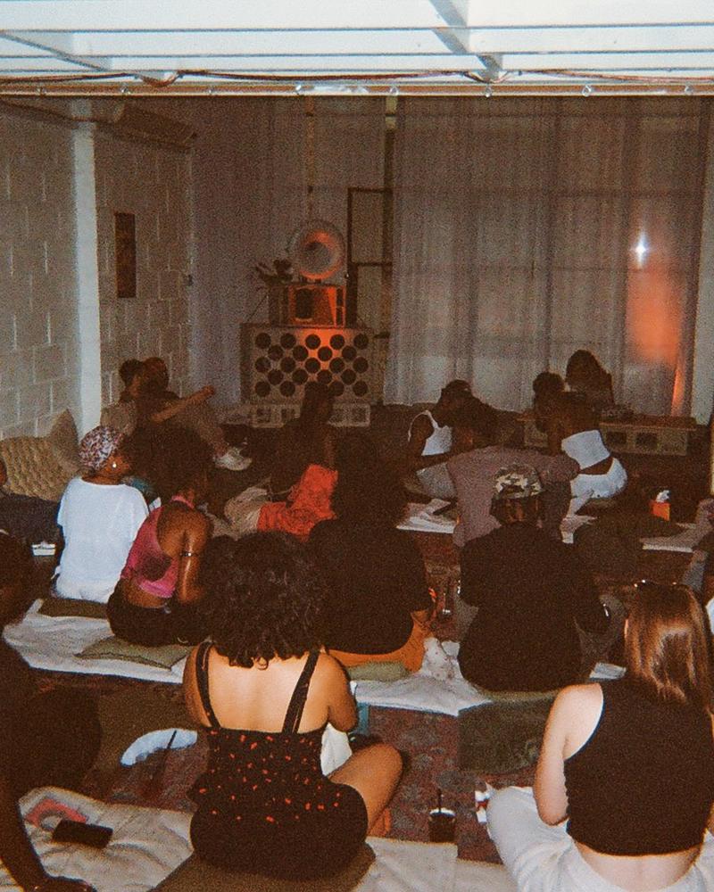 An audience seated on rugs facing the speakers
