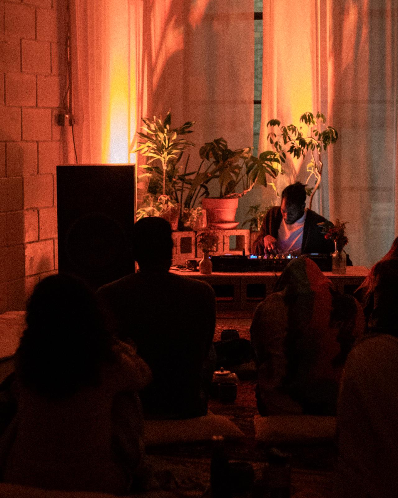 A DJ plays under warm orange light, surrounded by plants, with guests seated in front