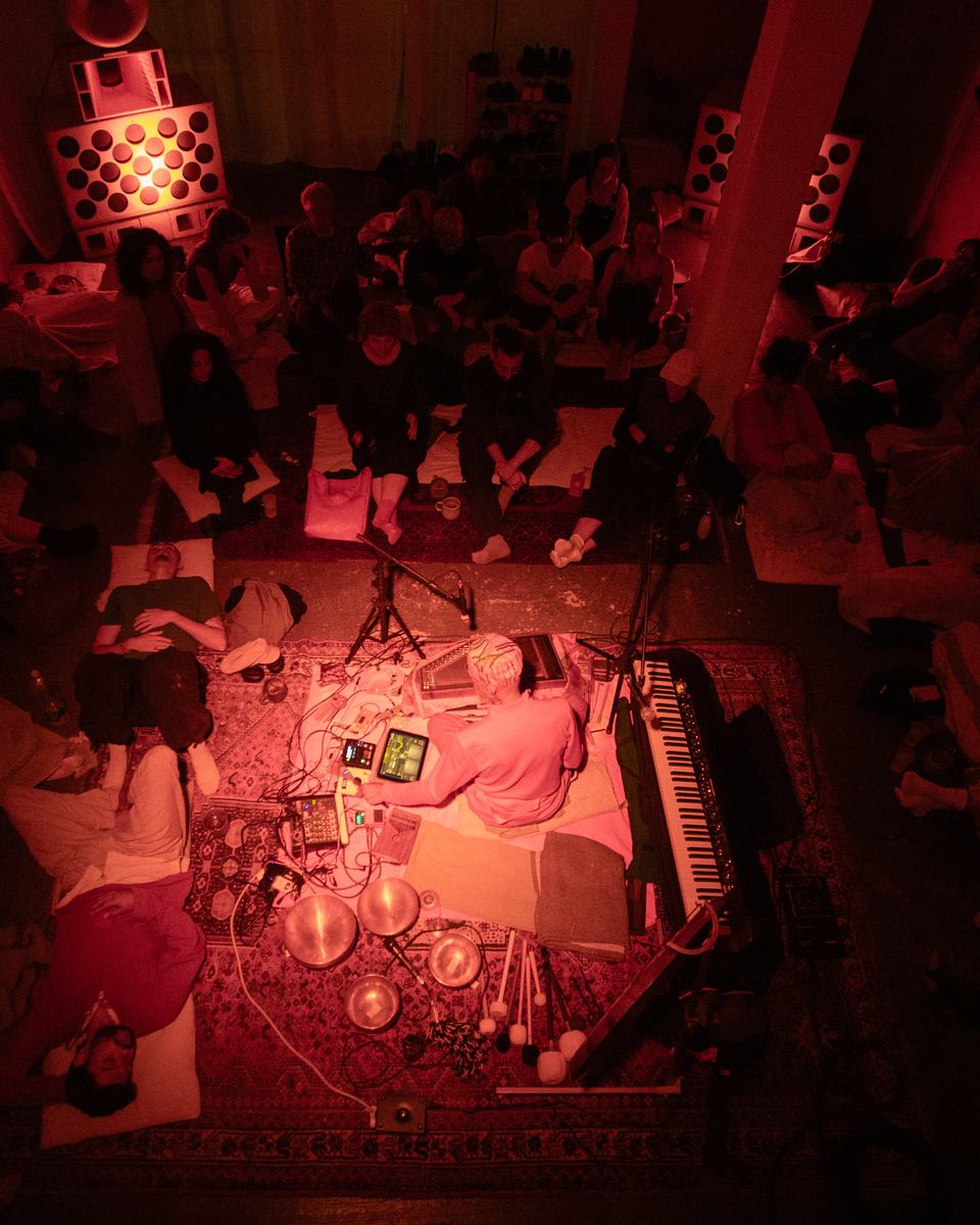 A musician surrounded by singing bowls, keys and electronics on a rug, with the audience lying and sitting around them